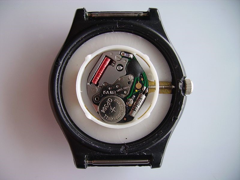 Basic quartz wristwatch movement. Bottom right: quartz crystal oscillator (silver tube), left bottom: cell watch battery (coin shape), top right: oscillator counter (in the black cover), top left: the coil of the stepper motor that powers the watch hands. Credit: https://en.wikipedia.org/wiki/Quartz_clock#/media/File:Armbanduhr_Rueckseite.jpg      